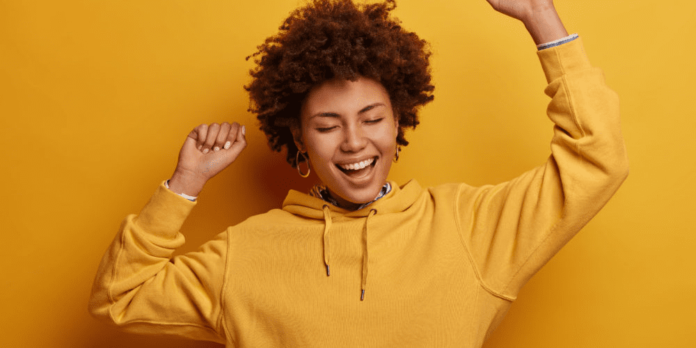 Woman in yellow hoodie in front of a yellow background smiling and holding hands in the air.