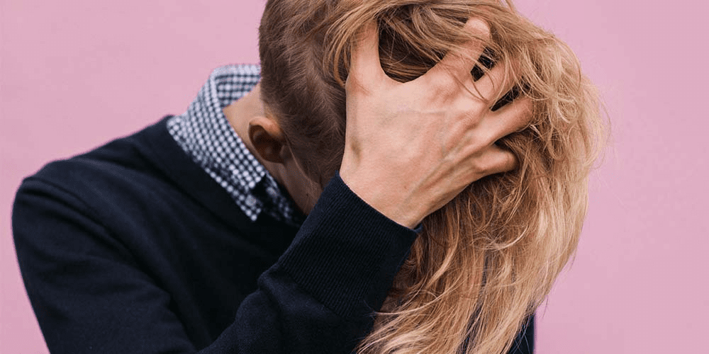 Person in front of a pink background looking down and holding head in hand.
