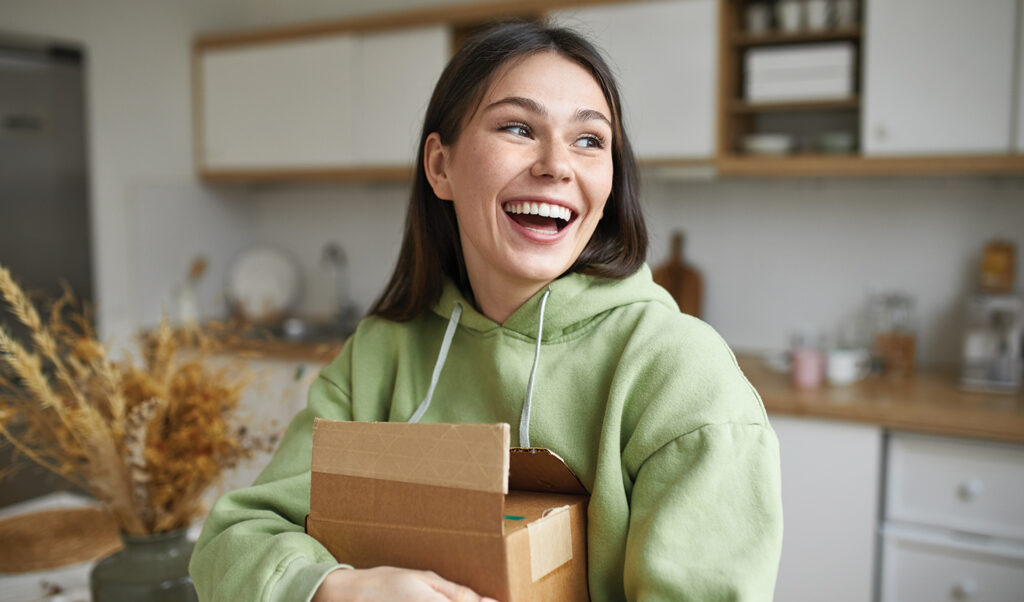A woman in a green hoodie, standing in a modern kitchen, holding a brown paper box and looking off camera, smiling. 