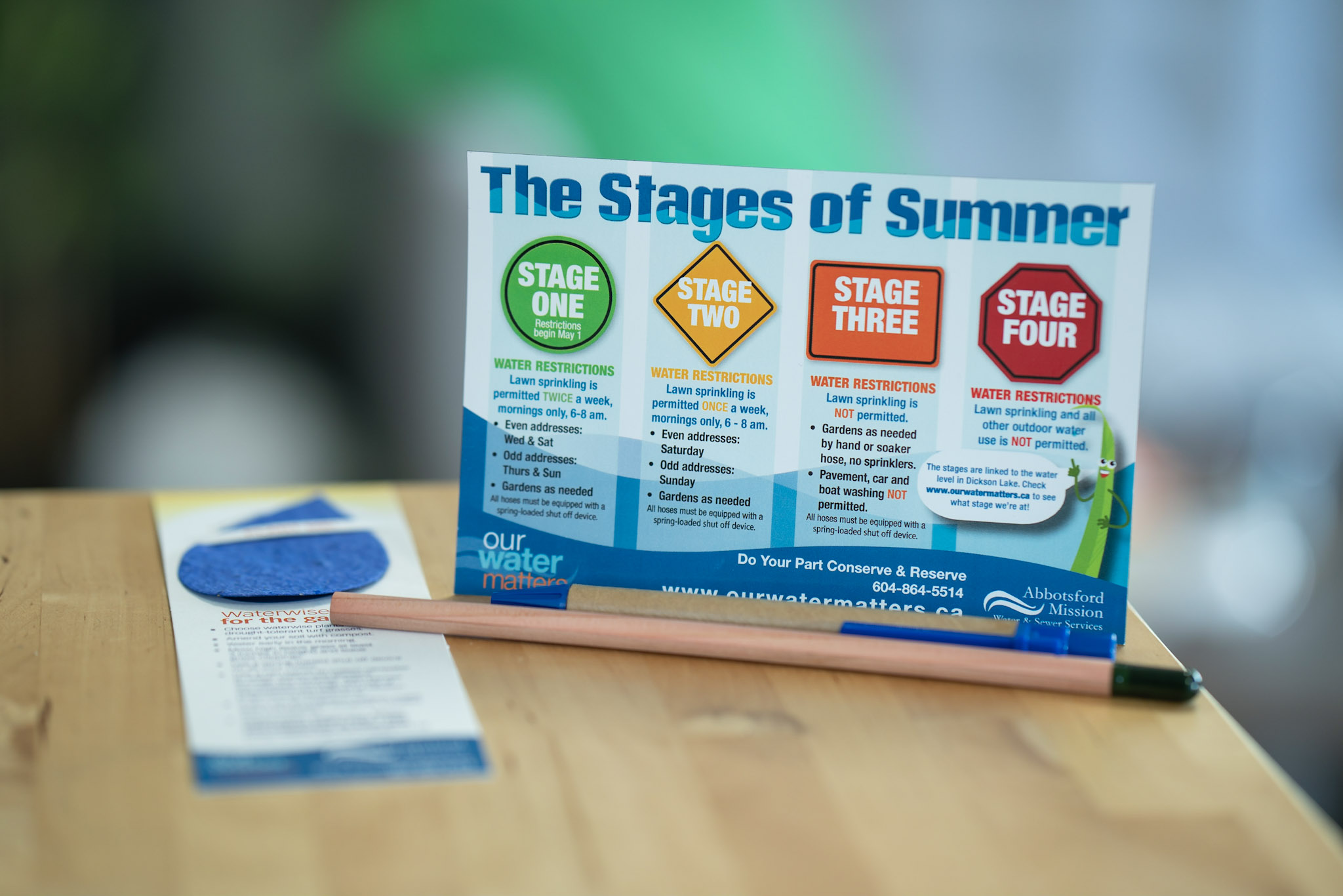 Image of a Post card with the stages of summer depicting stage one stage two stage three and stage 4 water restrictions for Abbotsford. Accompanied by a bookmark, pencil, and pen.