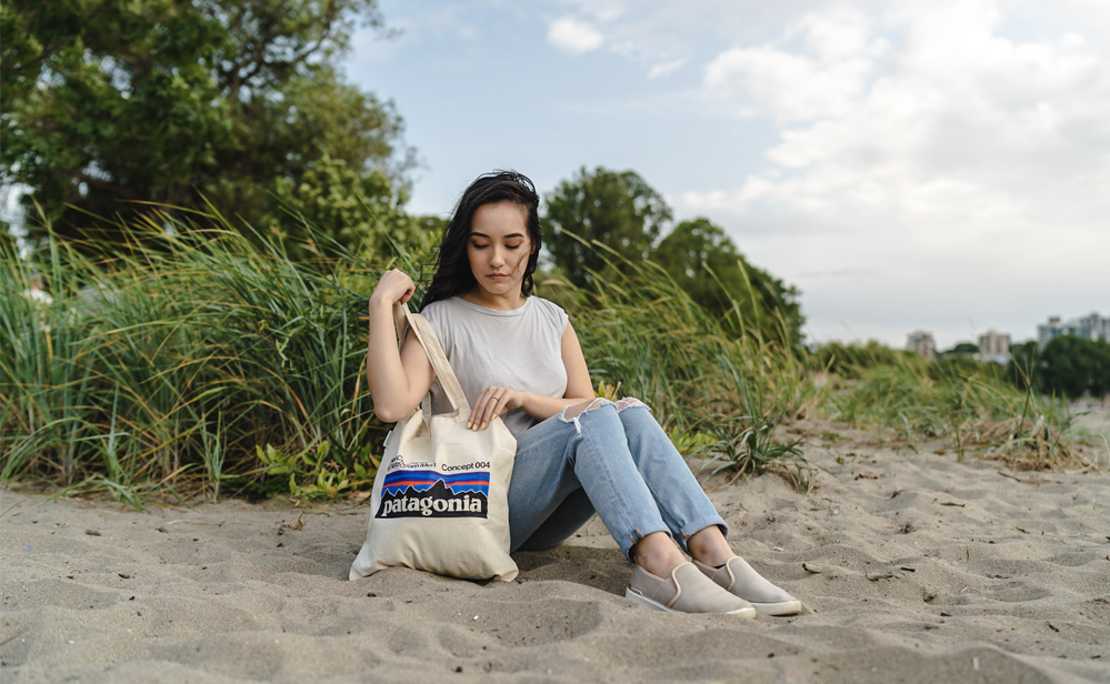 Person sitting on the beach in the sand holding a Patagonia branded cotton tote bag. Grass, trees and clouds are in the background.