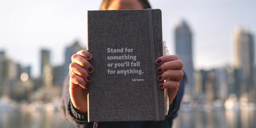 Person holding up grey notebook that says "stand for something or you'll fall for anything."