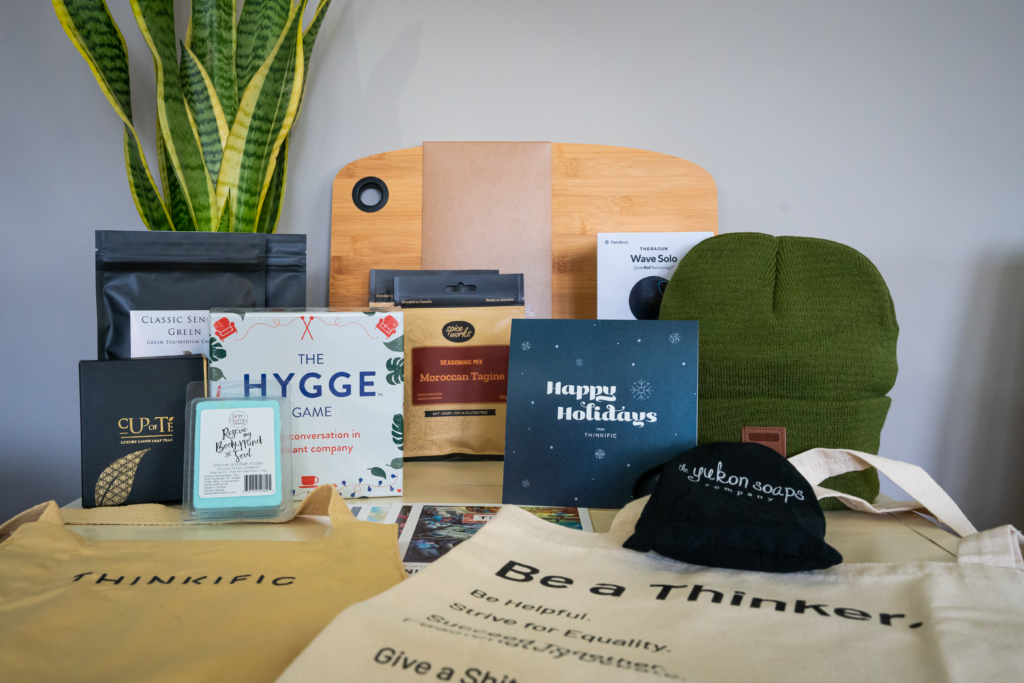 Thinkific branded tote bags and other gifts placed on a table in front of a green snake plant. Including a beanie, tea packages, soaps, games and a wooden cutting board.  