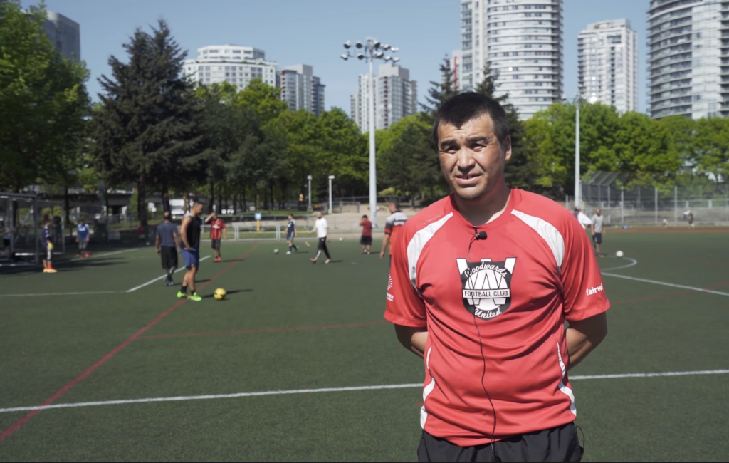 Participant of the Vancouver Street Soccer  League standing in a soccer field with people kicking a soccer ball in the background.