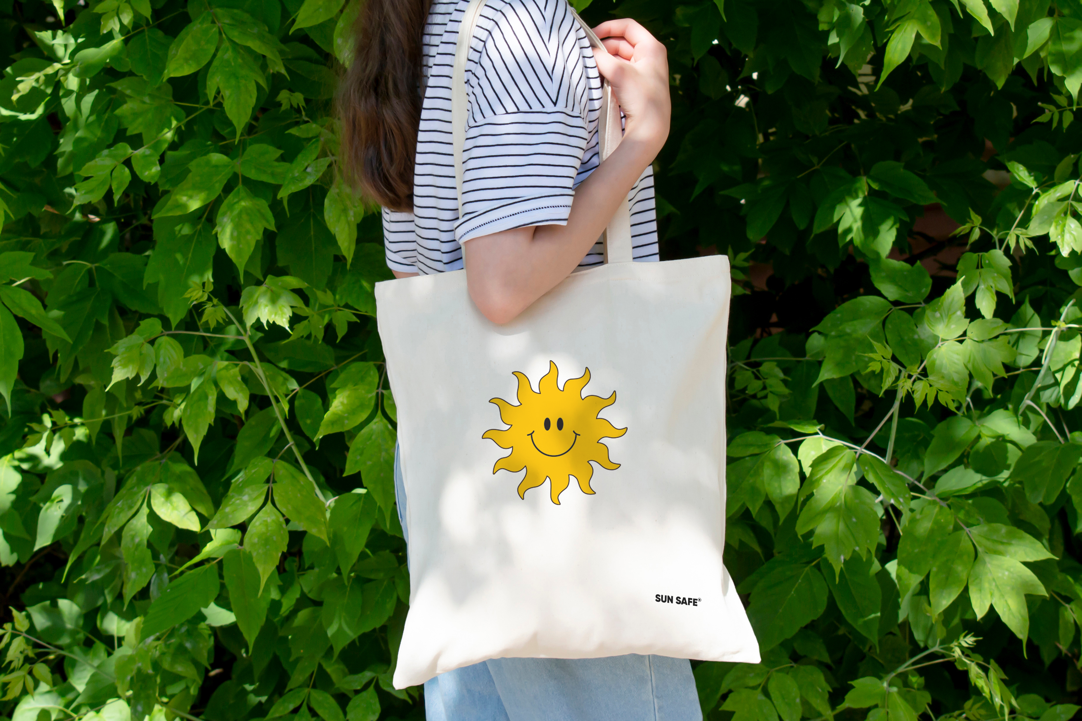 Person holding a tote bag with a graphic sun and the branded words sunsafe. Leafy green vines in the background.