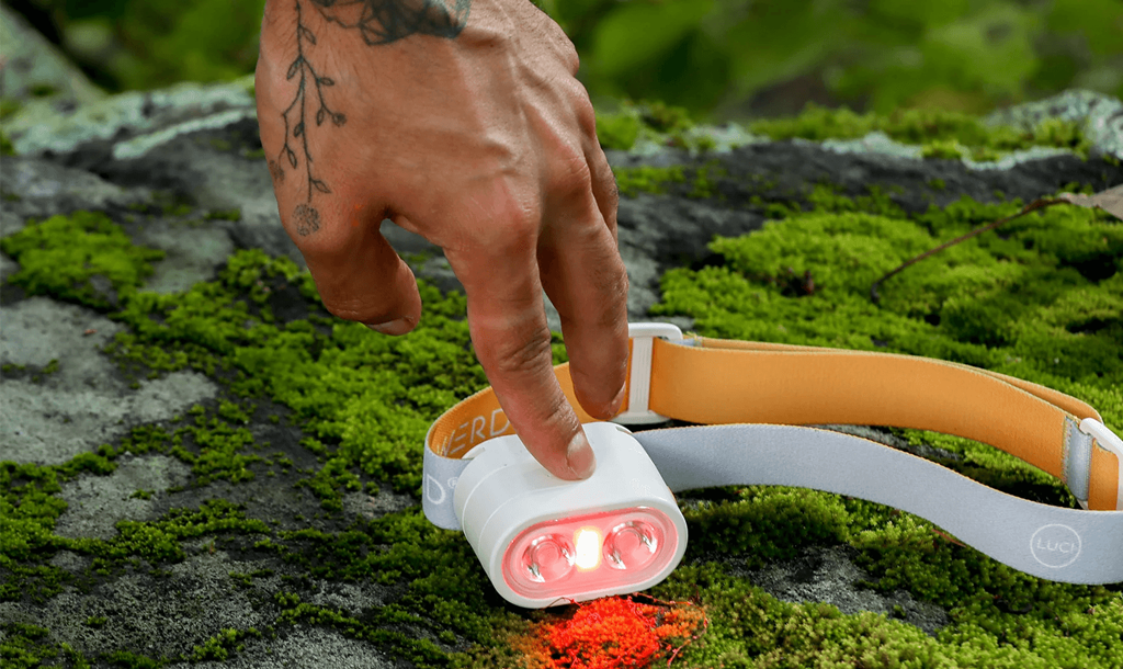 A person’s hand pressing a button on a headlamp sitting on a mossy green rock. 