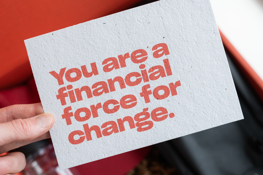 A person’s hand holding up a seed paper post card that reads “You are a financial force for change.”