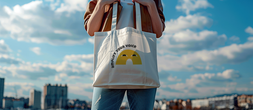 person holding a cotton totebag with graphic that reads amplify your voice and clouds in the background