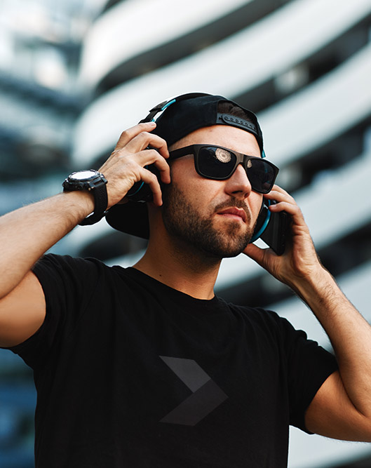 Portrait of a young bearded man with a black hat and sunglasses in black t-shirt with headphones in the city