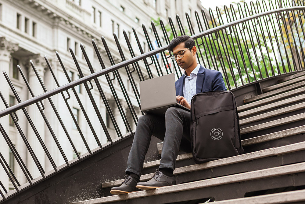 man sitting outside on stairs looking at his laptop wit h his bag made from recycled plastics sitting beside him.