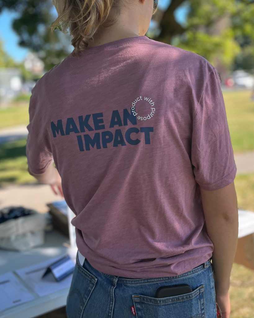 Woman wearing a pink t-shirt facing away. The back of the tee reads MAKE AN IMPACT - product with purpose.