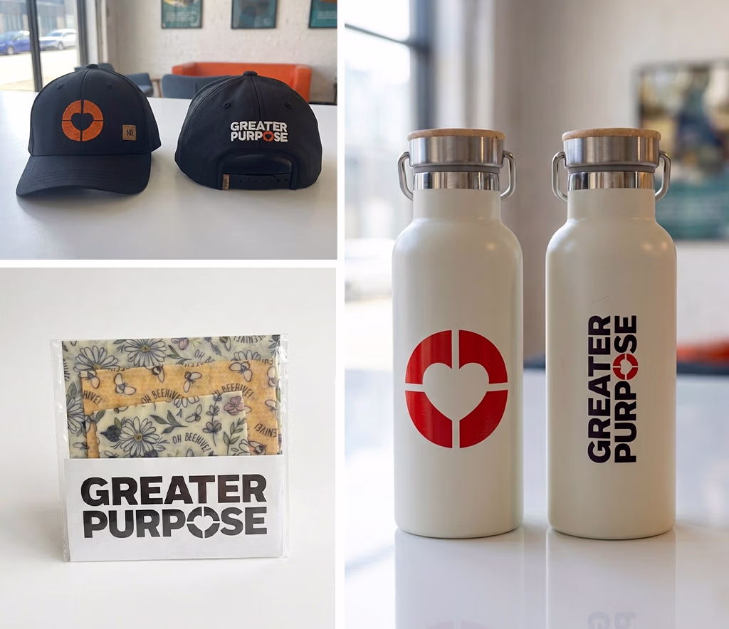 A collage showcasing branded promotional merchandise for Greater Purpose. The top left features the front and back of a black baseball cap with an orange circular logo on the front and "GREATER PURPOSE" embroidered in white on the back. The bottom left shows a reusable beeswax food wrap in branded packaging with the Greater Purpose logo. The right side displays two white insulated stainless steel water bottles with bamboo lids and metal handles—one printed with a red circular heart logo and the other with the vertical "GREATER PURPOSE" wordmark. The products are photographed on a white tabletop in a bright, modern office setting with a softly blurred background.