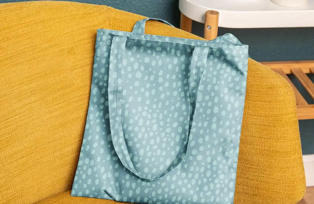 A light blue fabric tote bag covered in an all-over pattern of irregular light teal dots, resting on a textured yellow armchair. In the background, part of a white and wood side table is visible.